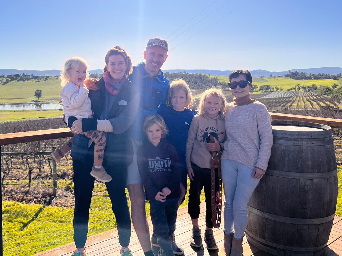 A family of seven posing together on a wooden deck overlooking a sunny vineyard landscape under a clear blue sky. A large wooden barrel is visible on the right side of the deck.